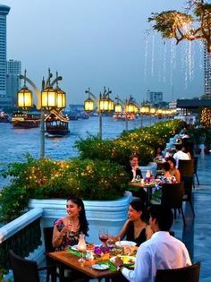 people are sitting at tables outside by the water with lights on them and boats in the background