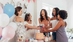 four women standing around a table with cake and drinks in front of balloons on the wall