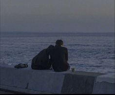 two people sitting on the edge of a concrete wall looking out to sea at dusk