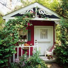 a small red and white house surrounded by greenery with a bench on the porch