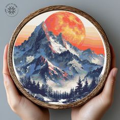 a hand holding up a cross - stitch picture of a mountain range with the sun setting