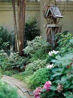 a garden with lots of plants and flowers around it in front of a house that has a birdhouse on the roof