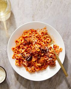 a white bowl filled with pasta and meatballs next to a glass of wine on a table