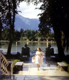 a woman in a white dress is walking down the sidewalk near some trees and water