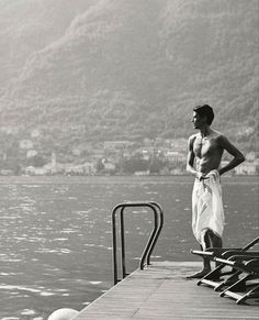 a shirtless man standing on a dock next to the water with his hands in his pockets