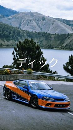 an orange and blue sports car driving down a road next to a lake with mountains in the background