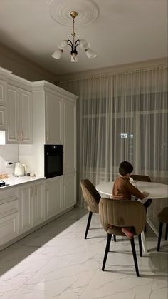 a person sitting at a table in a room with white cabinets and curtains on the windows