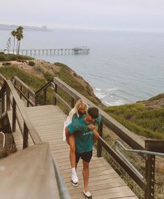 a man carrying a woman down a flight of stairs next to the ocean with a pier in the background