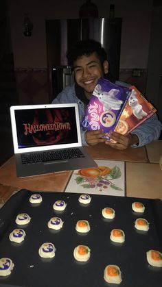 a young man holding up some halloween treats in front of a laptop and tray with mini cakes on it