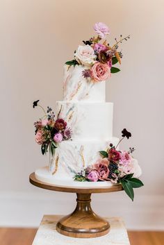 a white wedding cake with pink flowers and greenery on top is sitting on a gold pedestal