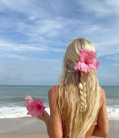 a woman with long blonde hair and pink flowers in her hair sitting on the beach