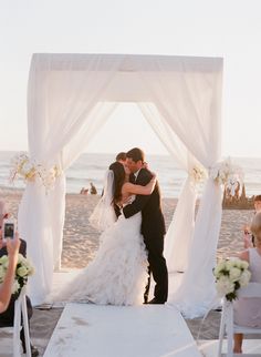 a bride and groom kissing on the beach in front of their wedding ceremony arch with white draping