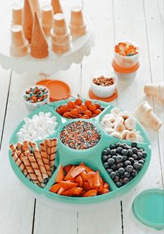 an assortment of food is arranged in a tray on a white wooden table, including carrots, pretzels, blueberries and crackers