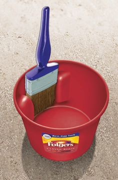 a red bucket with a blue brush in it on the ground next to some sand