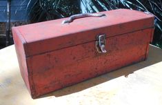 an old red box sitting on top of a table next to some plants and trees