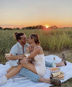 a man and woman sitting on a blanket holding wine glasses in front of the sun