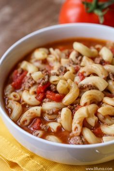 a white bowl filled with pasta soup next to a yellow napkin and tomatoes on the side