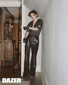 a man in black leather pants leaning against a wall with his hands on the door handle