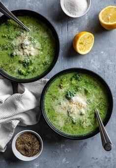 two bowls filled with broccoli soup next to some lemons and spices on a table