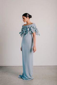 a woman in a long blue dress with flowers on the shoulder and shoulders, standing against a white wall