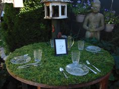 the table is set with plates and silverware on mossy ground next to a birdhouse