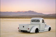 an old white truck parked in the middle of a desert area with mountains in the background