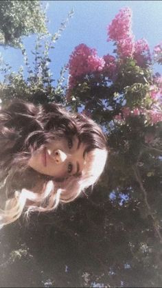 a woman with long hair standing in front of some trees and flowers looking up at the sky