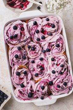 blueberry rolls in a white baking dish on a table with flowers and berries around them