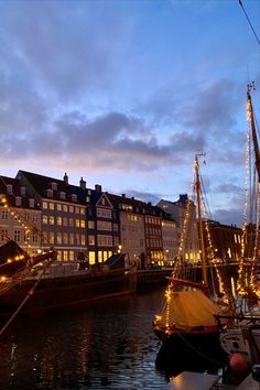 several sailboats are docked in the water near buildings and lights on them at dusk