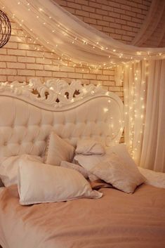 a white bed with lots of pillows and lights on the headboard, in front of a brick wall