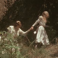 two women in white dresses holding hands and walking through tall grass with rocks behind them