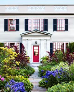 a white house with black shutters and red door surrounded by colorful flowers in front of it