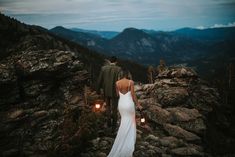 a bride and groom standing on top of a mountain looking at the view from their wedding dress