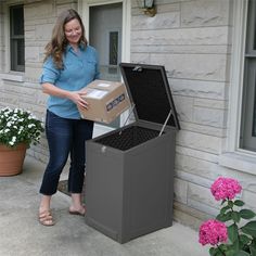 a woman holding a box and standing next to a trash can on the side of a house