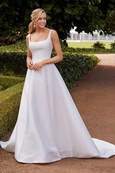 a woman wearing a white wedding dress standing in front of a tree and bushes with her hand on her hip