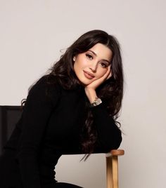 a woman sitting on top of a wooden chair next to a white wall with her hand under her chin