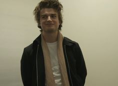 a young man wearing a black jacket and tan scarf standing in front of a white wall