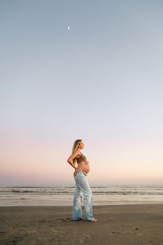 a pregnant woman standing on the beach at sunset with her belly exposed and hands behind her back