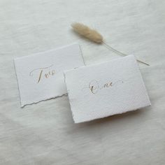 two white cards with gold calligraphy on them and a feather resting on the top