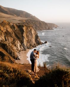 a man and woman standing on top of a cliff next to the ocean holding each other