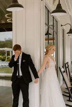 a bride and groom standing next to each other in front of a window holding hands