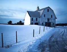 an old white barn in the middle of winter with snow on the ground and fence around it