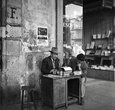 two men sitting at a desk in front of a book store, one reading the paper