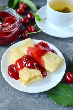 two crepes on a plate with cherries next to them and a cup of tea