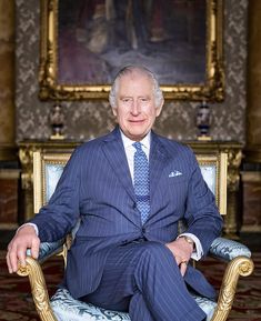 an older man in a blue suit and tie sitting on a chair with his legs crossed