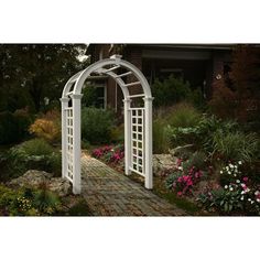 a white garden arch in the middle of a brick walkway surrounded by flowers and shrubs