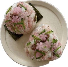 two pieces of sushi on a white plate with green garnish and pink flowers