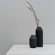 two black vases sitting on top of a white table next to each other and one has a plant in it