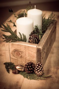 two white candles sitting on top of a wooden box filled with pine cones and greenery