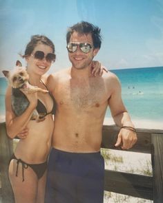 a man and woman pose for a photo on the beach with their dog in front of them